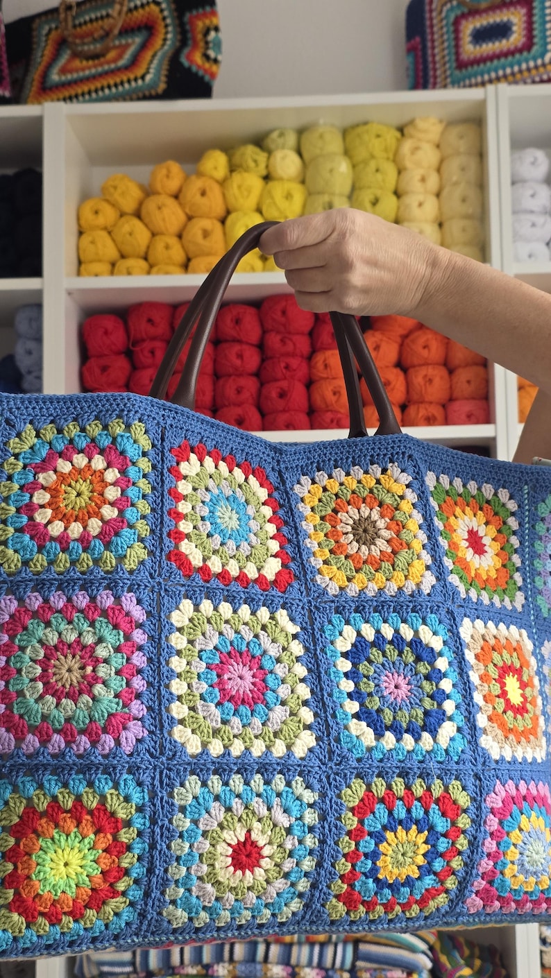 May include: A blue crocheted tote bag with a colorful granny square design. The bag features brown leather handles and is filled with a variety of yarn colors in the background.
