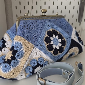 Könnte beinhalten: Handgefertigte Häkeltasche in Blau-, Weiß- und Beigetönen mit floralem Design. Die Tasche hat einen Metallverschluss im Vintage-Stil und einen hellblauen Schulterriemen. Die Clutch ist etwa 30 cm breit.