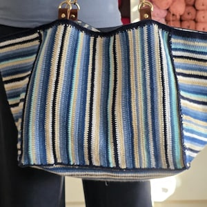 Könnte beinhalten: Eine große, handgefertigte Häkeltasche mit vertikalen Streifen in Blau-, Beige- und Weißtönen. Die Tasche hat braune Ledergriffe, die mit goldfarbener Hardware befestigt sind. Die Tasche wird von einer Person gehalten.