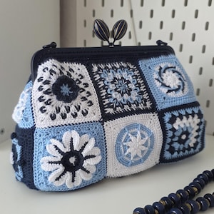 Può includere: Pochette all'uncinetto fatta a mano nei toni del blu e del bianco, con un design patchwork geometrico. La borsa ha una cornice e una chiusura blu scuro e include una tracolla con perline. La pochette è larga circa 25 cm.