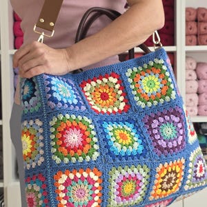 May include: A large, handmade blue crochet tote bag with colorful square patterns. The bag features a brown leather handle and a matching shoulder strap with gold-colored hardware. The bag is filled with a variety of colorful yarn.