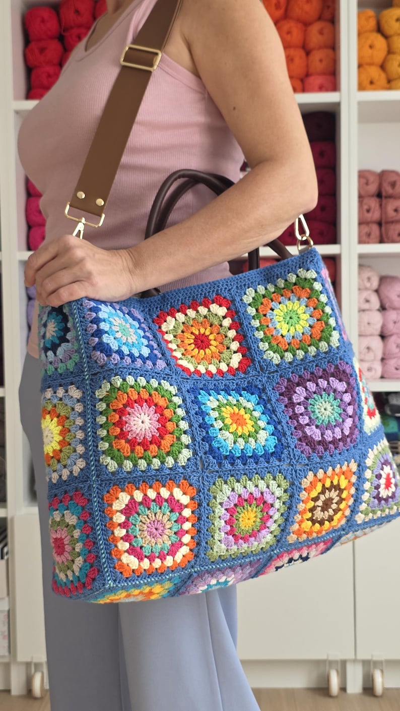 May include: A large, handmade blue crochet tote bag with colorful square patterns. The bag features a brown leather handle and a matching shoulder strap with gold-colored hardware. The bag is filled with a variety of colorful yarn.