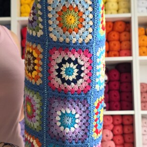 May include: A cylindrical, handmade crochet pillow in a vibrant granny square pattern. The pillow features a blue background with colorful squares in shades of yellow, orange, pink, purple, and blue. The pillow is held in front of a shelf filled with yarn.