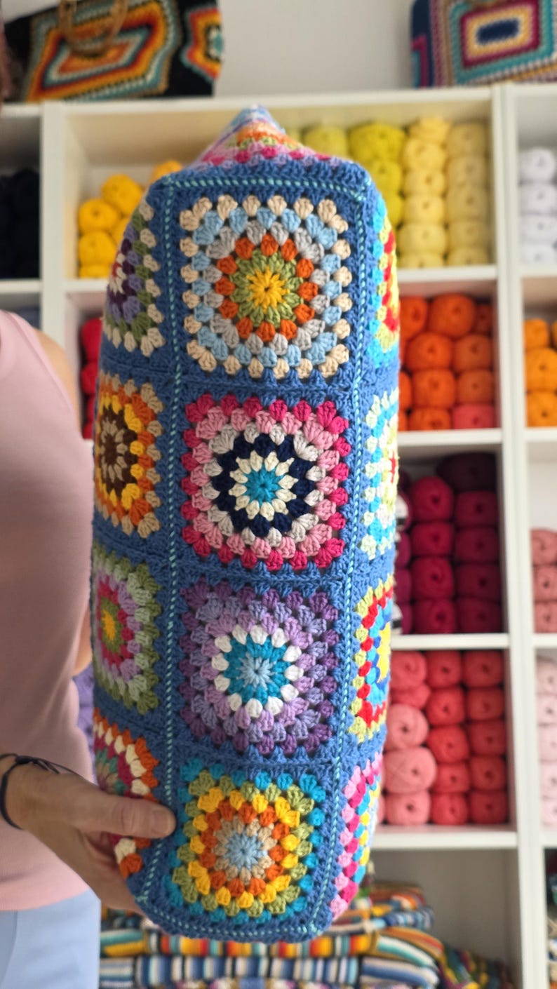 May include: A cylindrical, handmade crochet pillow in a vibrant granny square pattern. The pillow features a blue background with colorful squares in shades of yellow, orange, pink, purple, and blue. The pillow is held in front of a shelf filled with yarn.