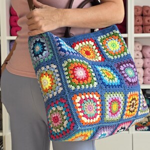 May include: A large, blue crochet tote bag with a colorful granny square design. The bag features a brown leather handle and strap. The squares are in shades of red, orange, yellow, green, blue, and purple.