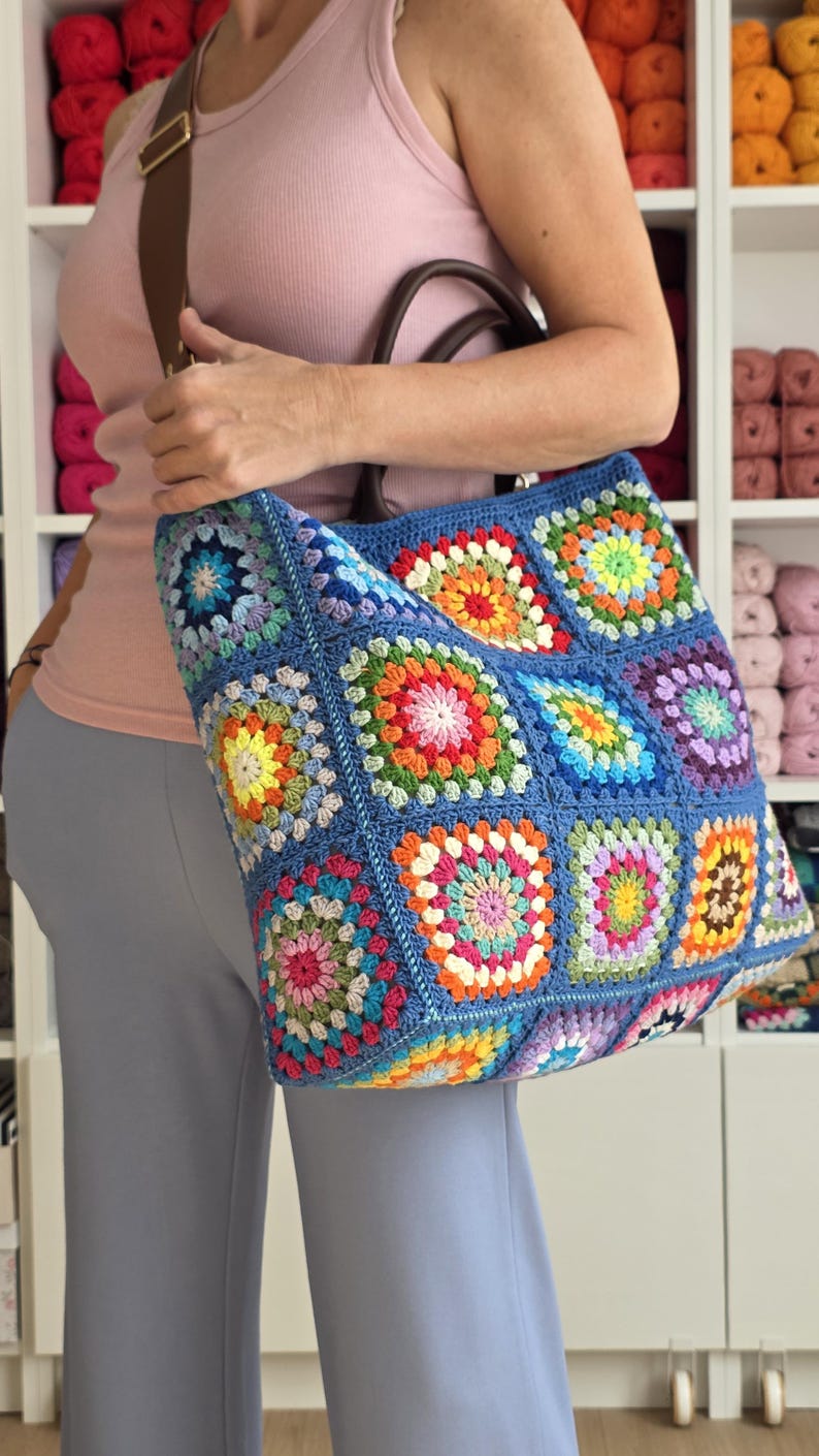 May include: A large, blue crochet tote bag with a colorful granny square design. The bag features a brown leather handle and strap. The squares are in shades of red, orange, yellow, green, blue, and purple.