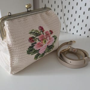 Könnte beinhalten: Cremefarbene Häkeltasche mit einem floralen Kreuzstichdesign in Rosa, Rot und Grün. Die Tasche hat einen Metallverschluss und einen abnehmbaren beigefarbenen Riemen. Die Tasche steht auf einer weißen Oberfläche.