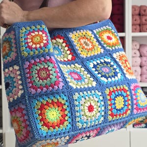 May include: A large, handmade tote bag with a blue background and colorful crochet squares. The squares feature vibrant patterns in red, yellow, green, and pink. The bag has a brown handle and is held by a person.