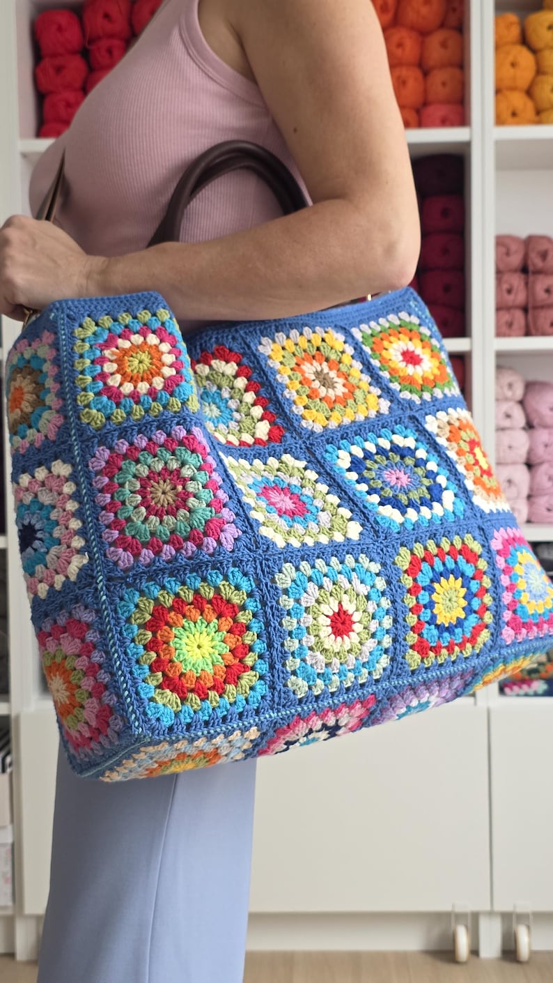 May include: A large, handmade tote bag with a blue background and colorful crochet squares. The squares feature vibrant patterns in red, yellow, green, and pink. The bag has a brown handle and is held by a person.