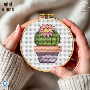 Puede incluir: Un bastidor de bordado de punto de cruz con un cactus de dibujos animados en una maceta con una cara sonriente. El cactus tiene una flor rosa y amarilla. El bastidor mide 10 cm de diámetro, con el texto "mini" encima de la medida.