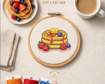 Pode incluir: Um bastidor de bordado em ponto cruz com um desenho de panquecas, incluindo manteiga, xarope e frutos silvestres. O bordado está num bastidor de madeira de 9,07 x 6,53 cm. A imagem também inclui um prato de panquecas, uma chávena de café e bobinas de linha.