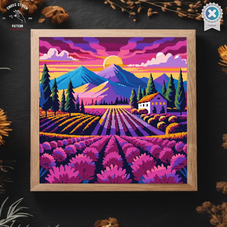 May include: A framed cross stitch pattern of a colorful landscape with a house, mountains, and lavender fields. The sky is a vibrant pink and orange with clouds. The house is white with a brown roof. The mountains are blue and purple. The lavender fields are a deep purple.