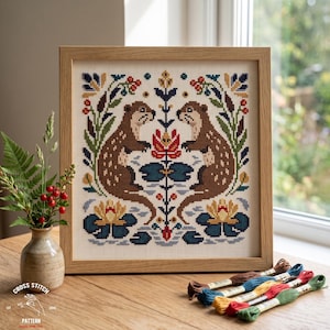 May include: A framed cross-stitch embroidery featuring two brown otters facing each other, surrounded by symmetrical floral motifs, red berries, and blue water lilies. The design is stitched on white fabric and housed in a light wood frame.