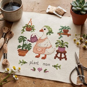 May include: Cross-stitch embroidery featuring a goose watering plants, surrounded by potted plants. The text "plant mom" is stitched below the goose. The design includes a coffee cup, thread, scissors, and a small succulent.