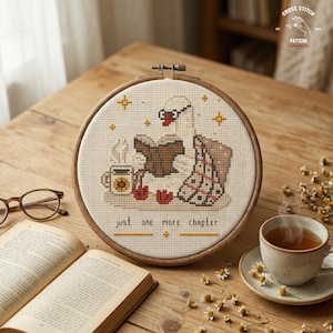 May include: A cross-stitch embroidery hoop featuring a swan reading a book, with a cup of tea and the text "just one more chapter." The design includes stars and a blanket. The hoop is set on a wooden table with a book and glasses.