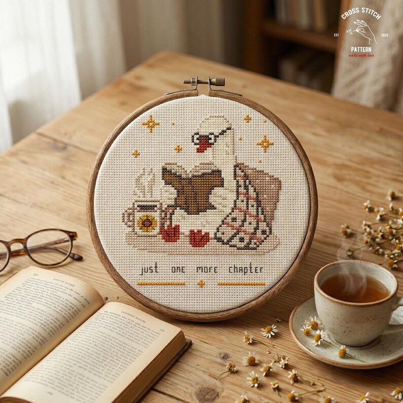 May include: A cross-stitch embroidery hoop featuring a swan reading a book, with a cup of tea and the text "just one more chapter." The design includes stars and a blanket. The hoop is set on a wooden table with a book and glasses.