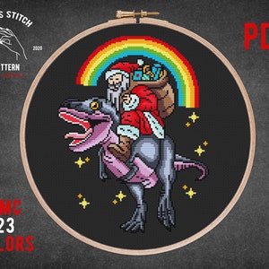 Puede incluir: Patrón de punto de cruz con Papá Noel montando un dinosaurio con un arcoíris detrás. El patrón está en un aro de bordado de madera e incluye 23 colores. El texto "PDF" está en la esquina superior derecha de la imagen.