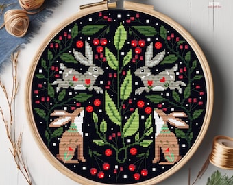 May include: Cross stitch pattern featuring two brown rabbits sitting under a green bush with red berries on a black background. The pattern is framed in a wooden hoop.