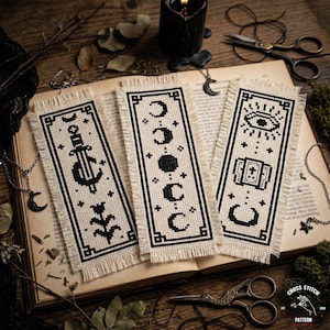 May include: Three black and white cross-stitch bookmarks with fringe, featuring a sword, moon phases, and an eye design. The bookmarks are on an open book with a candle, scissors, and other decorative items.