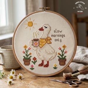 May include: Cross-stitch embroidery in a wooden hoop features a white goose with a yellow scarf, holding a coffee cup. The text "slow mornings only" is above the goose. The design includes a sun, flowers, and the words "Cross Stitch Pattern".