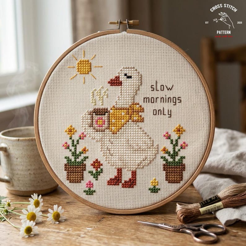 May include: Cross-stitch embroidery in a wooden hoop features a white goose with a yellow scarf, holding a coffee cup. The text "slow mornings only" is above the goose. The design includes a sun, flowers, and the words "Cross Stitch Pattern".
