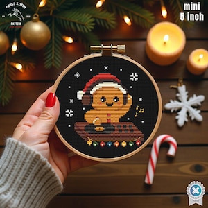May include: A mini 5-inch cross-stitch embroidery in a wooden hoop. The design features a gingerbread man wearing a Santa hat and headphones, with musical notes and snowflakes on a black background. The hoop is held by a person.
