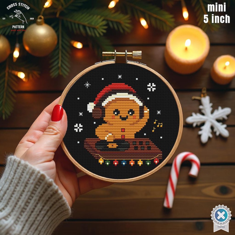 May include: A mini 5-inch cross-stitch embroidery in a wooden hoop. The design features a gingerbread man wearing a Santa hat and headphones, with musical notes and snowflakes on a black background. The hoop is held by a person.