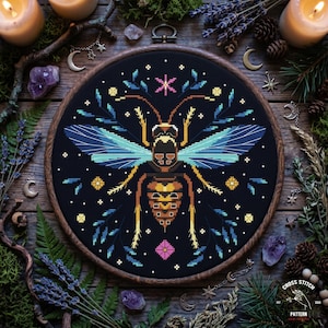 May include: A cross-stitch pattern of a bee with blue and yellow wings, set against a black background. The design is framed in a wooden hoop and surrounded by decorative elements like candles, crystals, and sprigs of greenery. The text "CROSS STITCH PATTERN" is visible.
