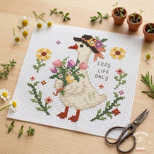 May include: Cross-stitch pattern featuring a goose wearing a hat adorned with flowers and holding a bouquet. The text "COZY LIFE ONLY" is displayed. The design includes floral elements and is set against a white background.