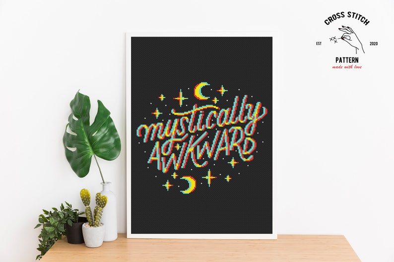 May include: Cross stitch pattern featuring the phrase "mystically awkward" in a retro, pixelated style with a rainbow color scheme. The design is surrounded by stars and crescent moons on a black background.