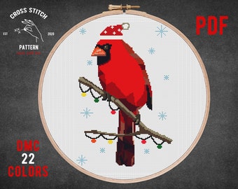 May include: Cross stitch pattern of a red cardinal wearing a Santa hat and perched on a branch with Christmas lights. The pattern is in a white embroidery hoop with a black background. The text "DMC 22 COLORS" is in the lower left corner of the image. The text "PDF" is in the upper right corner of the image.