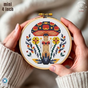 May include: A cross-stitch embroidery hoop featuring a colorful mushroom design. The mushroom has a red, orange, and blue cap, with yellow and blue flowers and foliage. The hoop is 4 inches (10 cm) in diameter and is held by hands.