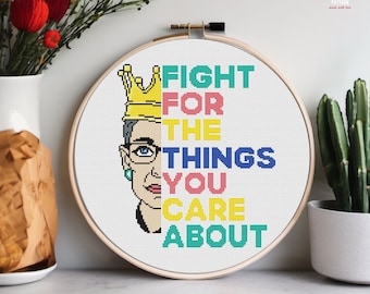 May include: A cross-stitch hoop featuring a portrait of a woman wearing glasses and a crown, with the text "FIGHT FOR THE THINGS YOU CARE ABOUT" in colorful letters. The hoop is displayed with a vase of flowers and a cactus.