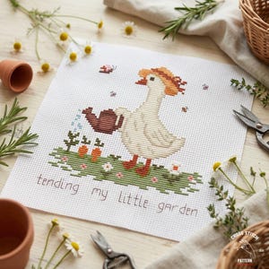 May include: Cross-stitch pattern featuring a goose wearing a straw hat, watering a garden with carrots and flowers. The text "tending my little garden" is stitched below the goose. The design is on white fabric.