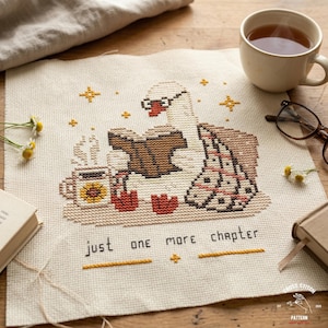 May include: Cross-stitch pattern featuring a swan wearing glasses and reading a book, with a cup of tea and the text "just one more chapter." The design includes stars and a blanket, all in a cozy, warm color palette.