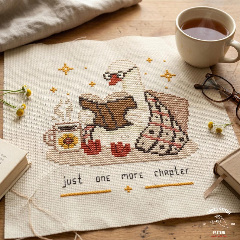 May include: Cross-stitch pattern featuring a swan wearing glasses and reading a book, with a cup of tea and the text "just one more chapter." The design includes stars and a blanket, all in a cozy, warm color palette.