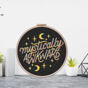 May include: A black embroidery hoop with a white background and the text "mystically awkward" in rainbow colors. The text is surrounded by small white stars and crescent moons.