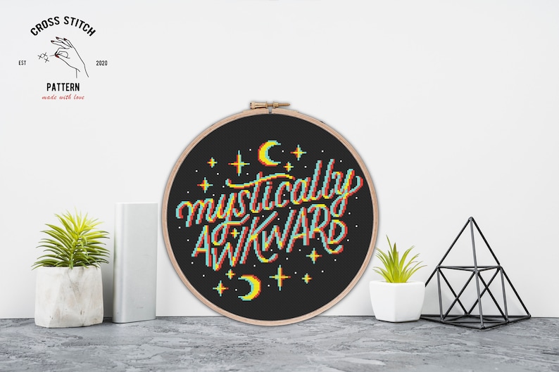 May include: A black embroidery hoop with a white background and the text "mystically awkward" in rainbow colors. The text is surrounded by small white stars and crescent moons.