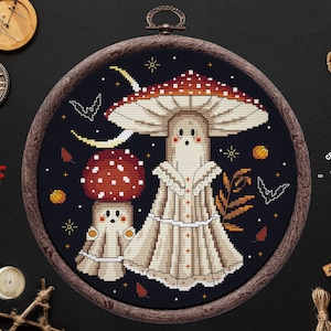 May include: Cross stitch pattern featuring two mushroom ghosts with red and white caps, a crescent moon, and bats on a dark blue background. The pattern is framed in a wooden hoop.