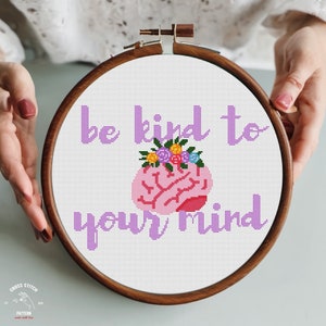 May include: A cross stitch pattern with a pink brain with flowers on top and the text "Be kind to your mind" in purple.
