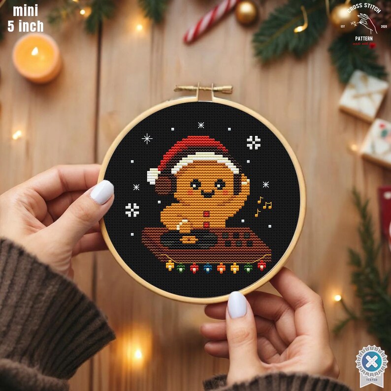 May include: A 5-inch diameter cross-stitch embroidery hoop featuring a gingerbread man wearing a Santa hat and headphones. The design includes snowflakes and musical notes on a black background. The hoop is held by hands.