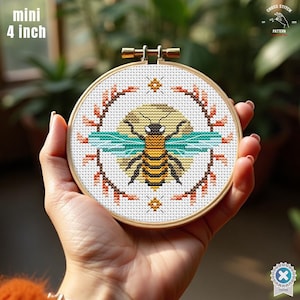 May include: A mini 4-inch cross-stitch embroidery hoop featuring a detailed bee design. The bee has yellow and black stripes, teal wings, and is surrounded by a floral border. The text "mini 4 inch" is visible.