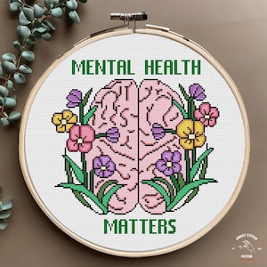May include: Cross stitch pattern featuring a pink brain with green leaves and colorful flowers. The text "Mental Health Matters" is stitched in green.