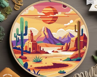 Può includere: Uno schema di punto croce che rappresenta un paesaggio desertico con un grande sole arancione, montagne viola, un fiume e due cactus. Lo schema è in un telaio da ricamo in legno.