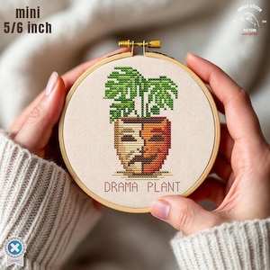 May include: A mini cross-stitch embroidery in a wooden hoop, measuring 5/6 inch. The design features a green plant in a pot shaped like a theater mask, with the text "DRAMA PLANT" below. The cross-stitch is on a beige fabric.