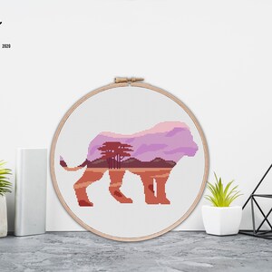 May include: Cross stitch pattern of a lion silhouette with a sunset landscape inside. The pattern is in a wooden embroidery hoop.