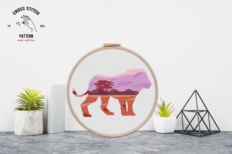 May include: Cross stitch pattern of a lion silhouette with a sunset landscape inside. The pattern is in a wooden embroidery hoop.