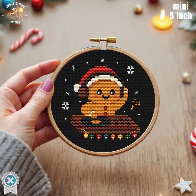 May include: A mini 5-inch cross-stitch pattern in a wooden hoop. The design features a gingerbread man wearing a Santa hat and headphones, sitting on a DJ setup with colorful lights. The background is black with white snowflakes.