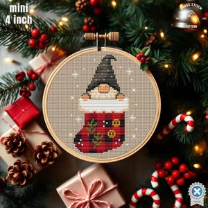 May include: A mini 10 cm cross-stitch embroidery hoop featuring a gnome in a Christmas stocking design. The gnome wears a black hat and peeks out from a red and black plaid stocking. The hoop is surrounded by festive holiday decorations.