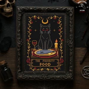 May include: Cross-stitch art in a black ornate frame. The design features a black cat with red eyes, a crescent moon, and the text "THE PRIORITY FOOD." A glass of wine, a candle, and a fish on a plate are also depicted.